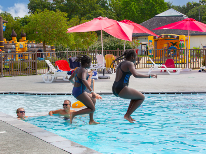 Two kids jump into the pool at Flower Camping L'Ile d'Offard, with loungers and red umbrellas beside them.