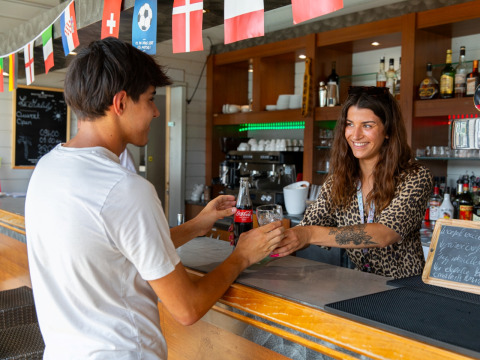 Een lachende barmedewerkster serveert een drankje aan een gast bij Flower Camping L'Ile d'Offard, Pays de la Loire.