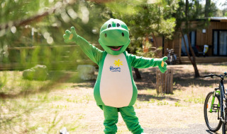 Person in a green dinosaur costume standing outdoors at a glamping holiday park with cabins and bikes.