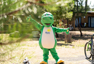 Persona disfrazada de dinosaurio verde en un parque de glamping con bicicletas y cabañas de fondo.