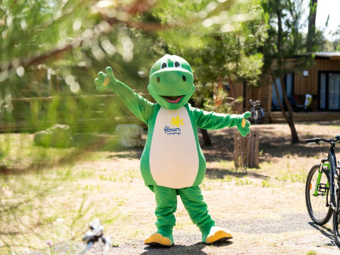 Personne en costume de dinosaure vert debout dans un parc de glamping avec cabanes et vélos autour.
