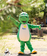 Person in a green dinosaur costume standing outdoors at a glamping holiday park with cabins and bikes.
