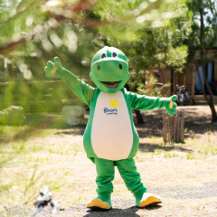 Eine Person im grünen Dinosaurierkostüm steht in einem Glamping-Park mit Fahrrädern und Holzhütten.