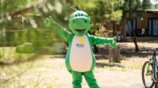 Eine Person im grünen Dinosaurierkostüm steht in einem Glamping-Park mit Fahrrädern und Holzhütten.