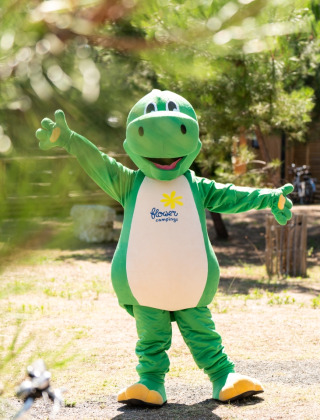 Person in a green dinosaur costume standing outdoors at a glamping holiday park with cabins and bikes.
