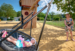 Een kind ligt ontspannen op een nestschommel terwijl een vrouw toekijkt op de speeltuin van Flower Camping L'Ile d'Offard.