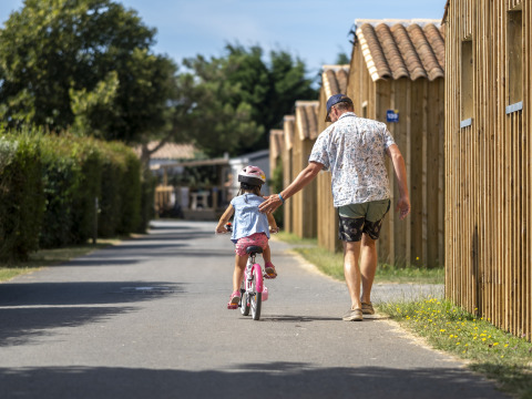 Un hombre ayuda a un niño en bicicleta en un camino, junto a cabañas de madera en un sitio de glamping.