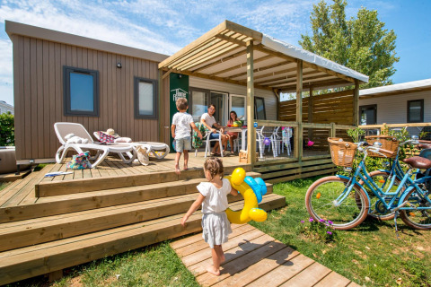 Familie nyder sommerferie på træterrasse ved Flower Camping Les Ilates, med børn og cykler i forgrunden.