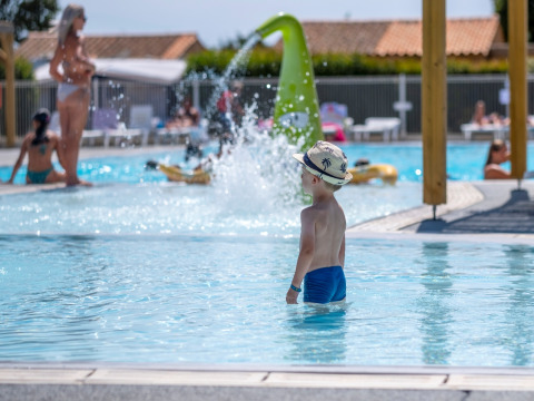 Un bambino con cappello si trova nella piscina di Flower Camping Les Ilates, parco vacanze in Nouvelle-Aquitaine, Francia.