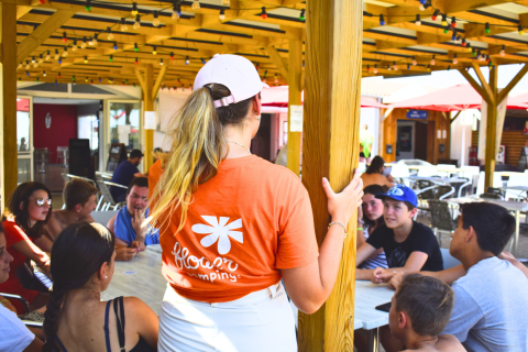 Jugendliche sitzen gemeinsam an einem Tisch unter einer überdachten Terrasse bei Flower Camping Les Ilates.