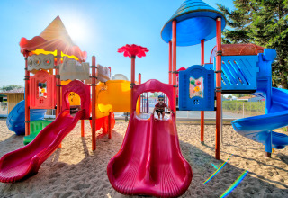 Farverigt legepladsområde med rutsjebaner og sand, børn leger i solen på Flower Camping Les Ilates.