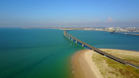 Luftfoto af en bro, der strækker sig over blåt vand ved et feriepark med glamping faciliteter på kysten.