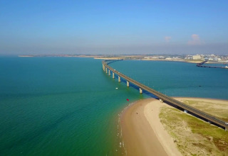 Vista aerea di un ponte che attraversa l’acqua blu accanto a un villaggio vacanze con glamping sul litorale.