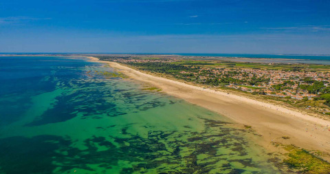 Vue aérienne d’un parc de vacances avec hébergements glamping, plage de sable et eau claire sous ciel bleu.