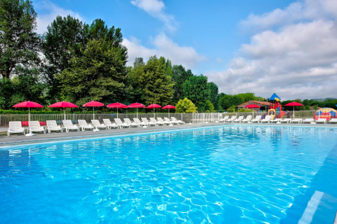 Piscina all'aperto con lettini bianchi e ombrelloni rossi, immersa nel verde di un villaggio turistico francese.