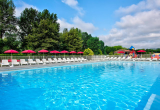 Piscina all'aperto con lettini bianchi e ombrelloni rossi, immersa nel verde di un villaggio turistico francese.