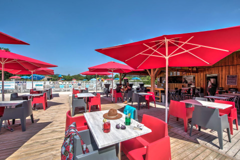 Terraza al aire libre en Flower Camping Les Ondines con sombrillas rojas, sillas de colores y piscina en Occitania, Francia.