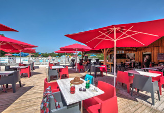 Udendørs terrasse på Flower Camping Les Ondines med røde parasoller, farverige stole og pool i Occitanie, Frankrig.