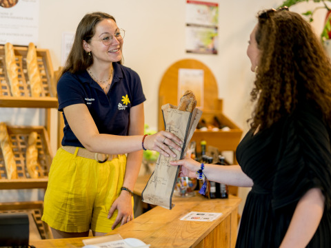 Eine Mitarbeiterin im Flower Camping Les Ondines überreicht einer Kundin ein Baguette an der Rezeption.
