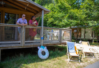 Two people relax on a wooden cabin porch with deck chairs and a float in a sunny holiday park setting.