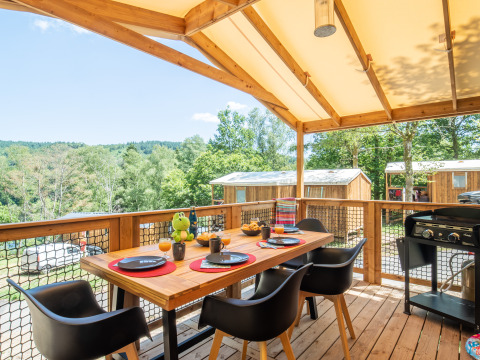 Covered wooden terrace with dining table and chairs at Flower Camping La Plage, overlooking cabins and greenery.