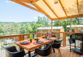 Covered wooden terrace with dining table and chairs at Flower Camping La Plage, overlooking cabins and greenery.