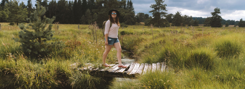 Mujer con gafas de sol y sombrero cruza un puente de madera en Flower Camping La Plage, Nouvelle-Aquitaine, Francia.