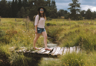 Femme en short, chapeau et lunettes traverse un pont en bois à Flower Camping La Plage, Nouvelle-Aquitaine, France.