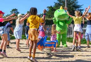 Børn leger og hopper i sandet med en stor, grøn maskot på en legeplads i Flower Camping Cap Finistère.