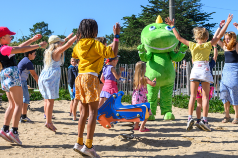 Des enfants jouent et sautent dans le sable avec une grande mascotte verte au Flower Camping Cap Finistère.