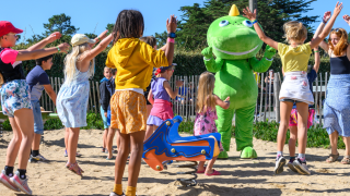 Des enfants jouent et sautent dans le sable avec une grande mascotte verte au Flower Camping Cap Finistère.
