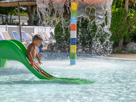 Un enfant glisse sur un toboggan vert dans une pataugeoire avec fontaine dans un parc de vacances en Bretagne, France.