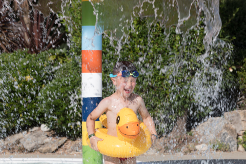 Kind met eend-zwemband en duikbril onder waterval in waterpark bij Flower Camping Cap Finistère, Bretagne.