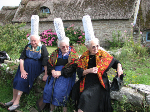 Drie oudere vrouwen in traditionele Bretonse kleding en hoge kanten mutsen zitten buiten op een stenen muur in Bretagne.