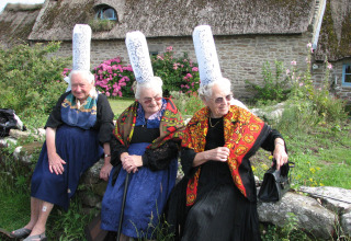 Tres mujeres mayores con trajes bretones tradicionales y altos sombreros de encaje sentadas en un muro de piedra en Bretaña.