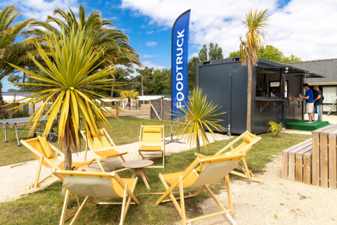 Outdoor food truck area at Flower Camping Le Vieux Moulin in Brittany, France, with palm trees and deck chairs.