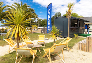 Udendørs foodtruck-område på Flower Camping Le Vieux Moulin i Bretagne, Frankrig, med palmer og stole.
