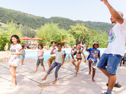 Kinderen dansen bij het zwembad met een begeleider bij Flower Camping Mas de Champel, omringd door heuvels.
