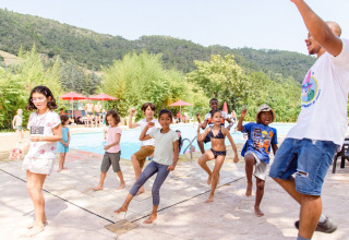 Des enfants dansent avec un animateur près de la piscine au Flower Camping Mas de Champel, entouré de montagnes.