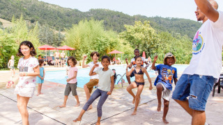Des enfants dansent avec un animateur près de la piscine au Flower Camping Mas de Champel, entouré de montagnes.