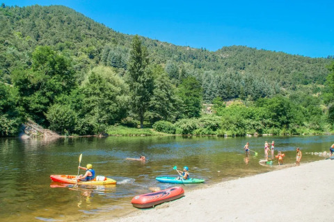 Kinder spielen und paddeln in Kajaks an einem Fluss inmitten grüner Hügel im Flower Camping Mas de Champel, Frankreich.