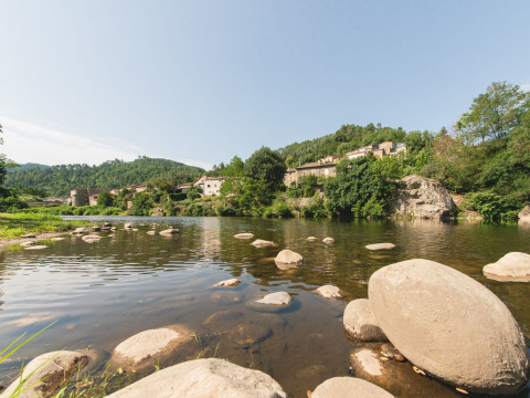 Flod med sten og udsigt til små huse og grønne bakker ved Flower Camping Mas de Champel i Auvergne-Rhône-Alpes.