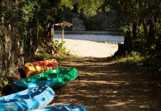 Kleurrijke kajaks liggen op een schaduwrijk pad dat naar een zandstrand bij Flower Camping Mas de Champel leidt.