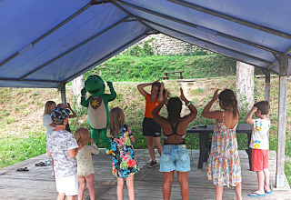 Kinder nehmen an einer Aktivität unter einem Pavillon mit einem als Dinosaurier verkleideten Maskottchen im Flower Camping Mas de Champel teil.
