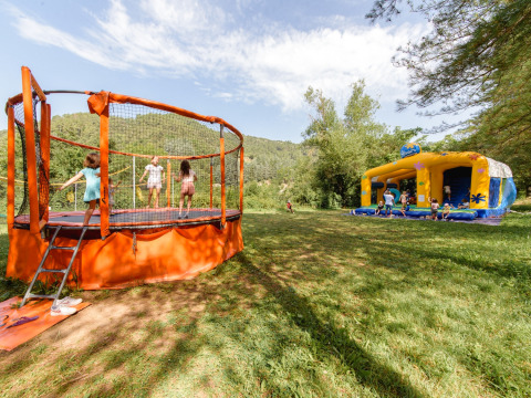 Børn leger på en trampolin og i en hoppeborg i grønne omgivelser på Flower Camping Mas de Champel i Frankrig.