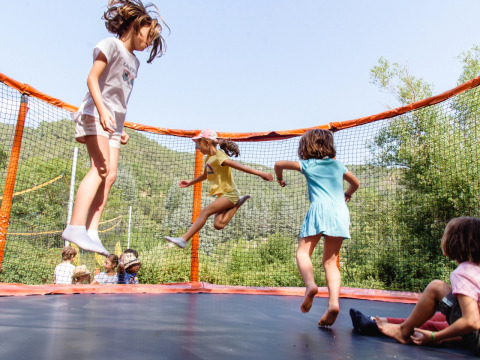 Niños jugando y saltando en una cama elástica con red en un parque vacacional rodeado de colinas verdes.