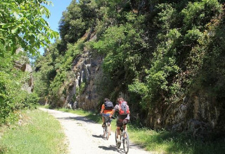 Two cyclists ride along a scenic path near Les Ollières sur Eyrieux in Auvergne-Rhône-Alpes, France, surrounded by greenery.