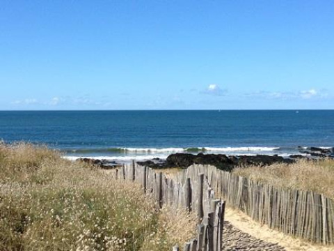 A sandy path with wooden fencing leads through grassy dunes to the sea at Flower Camping Les Paludiers, France.