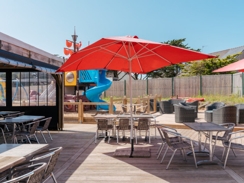 Terraza al aire libre con sombrillas rojas, mesas para comer y parque infantil en Flower Camping Les Paludiers, Pays de la Loire.
