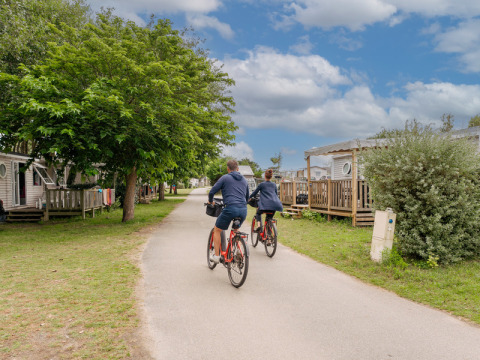 Dos personas montan en bicicleta en Flower Camping Les Paludiers, rodeados de casas móviles y vegetación.